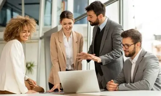 A group of four office workers collaborating around a laptop in a bright, modern office space.