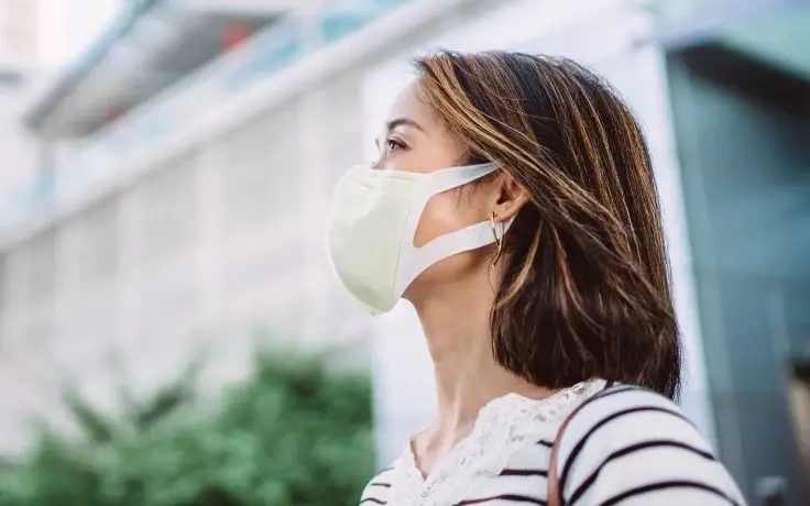 A woman with shoulder-length brown hair wears a white face mask and a striped shirt while looking up outdoors.