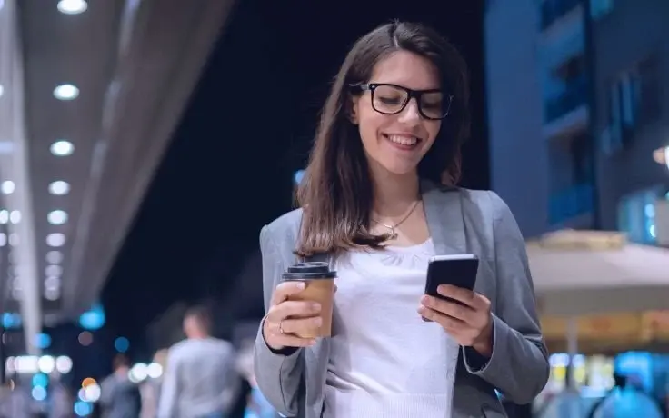 A smiling woman wearing glasses and a gray blazer holds a coffee cup and looks at her smartphone on a city street at night.
