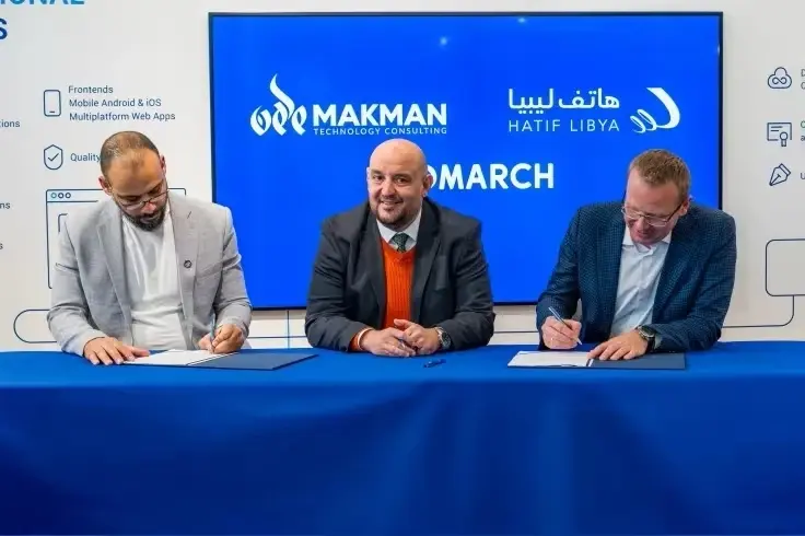 Three men in suits are at a table; the outer two are signing documents, while the central man smiles at the camera.