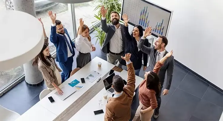 An overhead view of a group of business professionals celebrating with hands raised in a bright, modern office conference room.