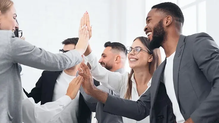 A diverse group of six colleagues, viewed from a low angle, give a celebratory high-five together in a bright office.