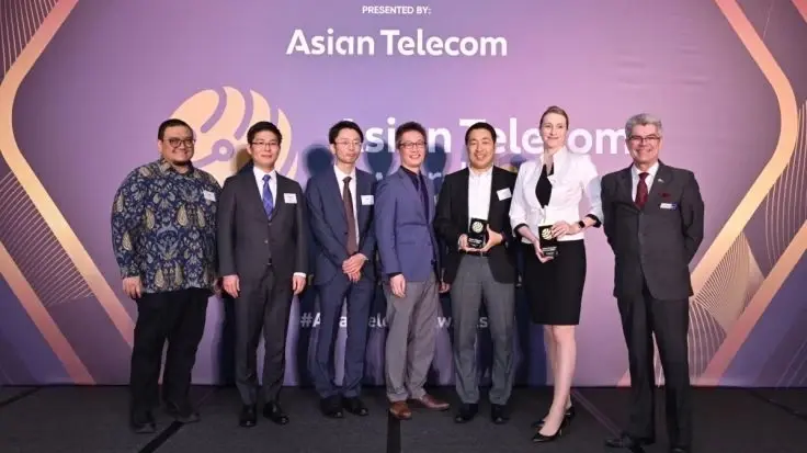 A group of professionals posing for a photo at an Asian Telecom Awards ceremony, with several members holding trophies in front of a purple backdrop.