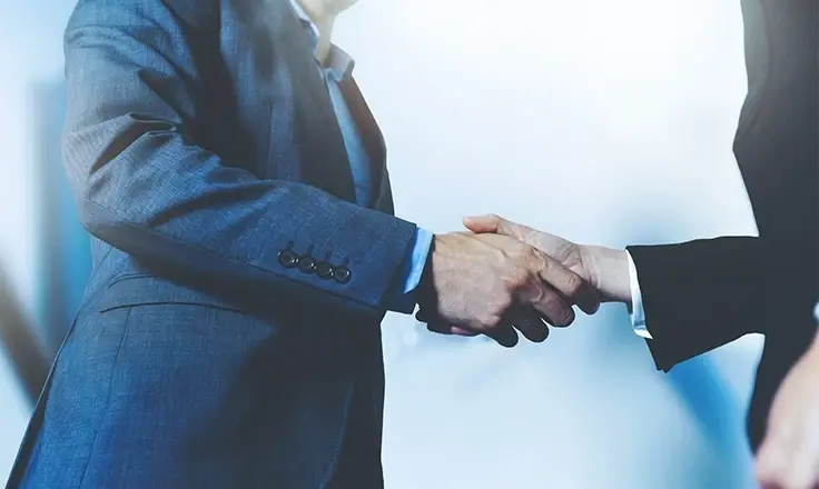 A close-up shot of two business professionals in suits shaking hands in a brightly lit environment.