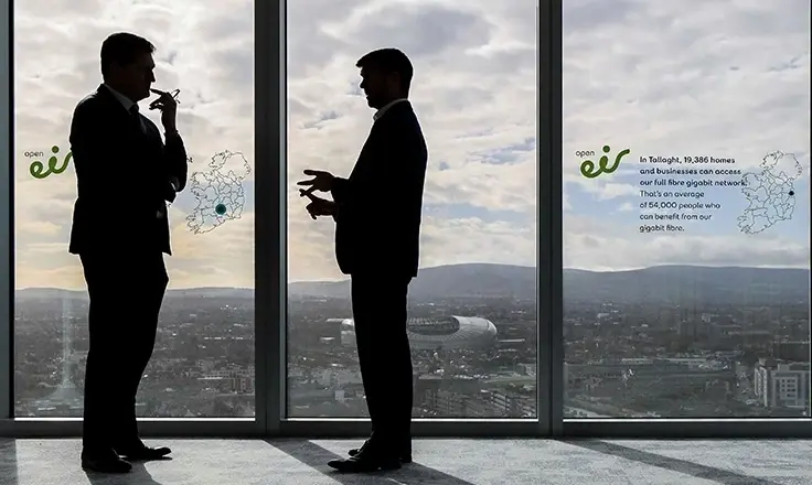 The silhouettes of two men in suits standing in front of a large window overlooking a city, with "open eir" branding on the glass.