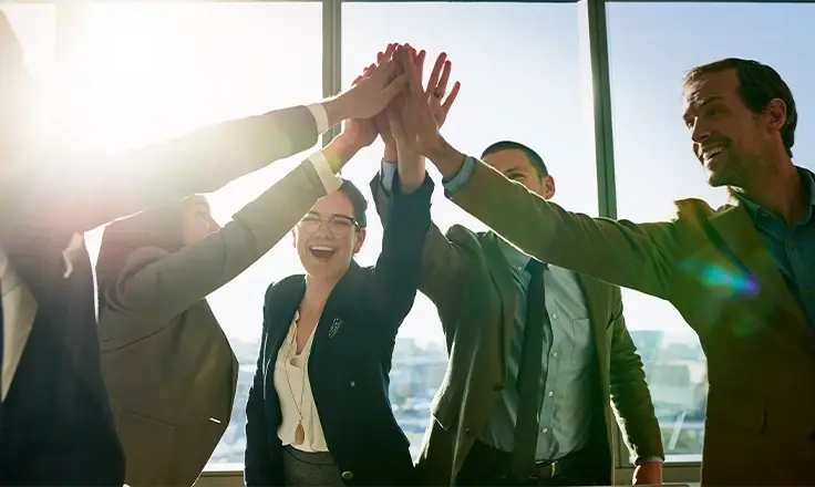 A business team of five celebrates with a stacked hand high five in a bright, modern office with city views.
