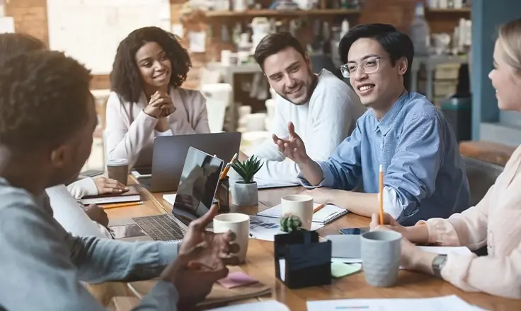 A group of young office workers sitting around a table, smiling and engaged in a discussion during a meeting in a modern office space.