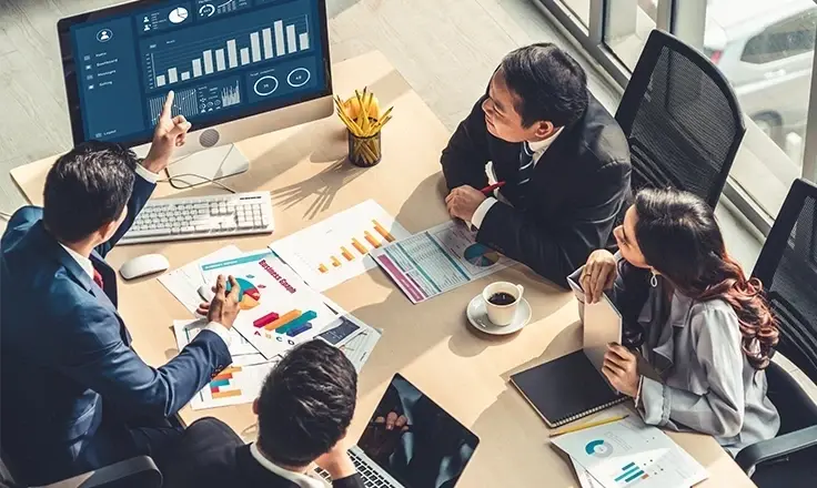 A team of professionals in a meeting, analyzing data and charts on a computer screen and printed documents spread across a desk.