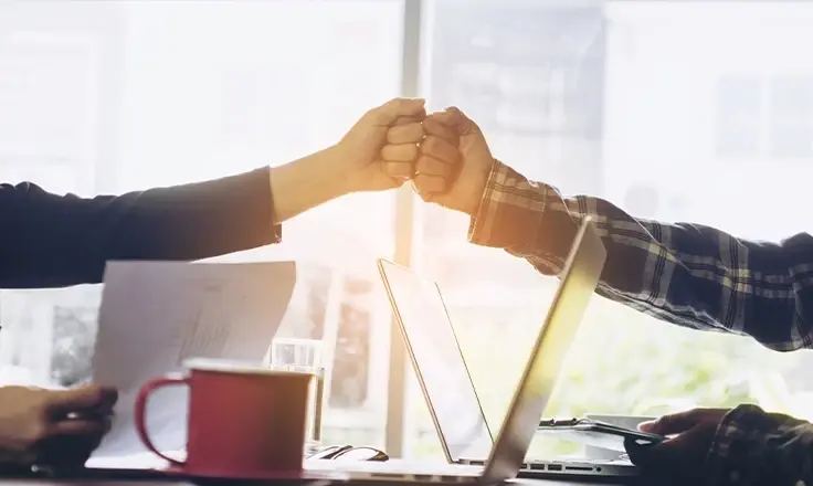 A close-up of two people giving a fist bump over an office desk with two laptops, papers, and a red mug.