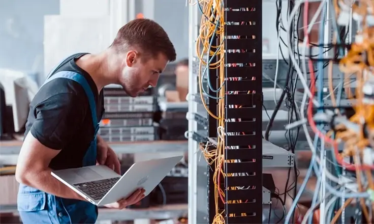 An IT technician holding a laptop while inspecting complex network cabling and server racks in a data center.