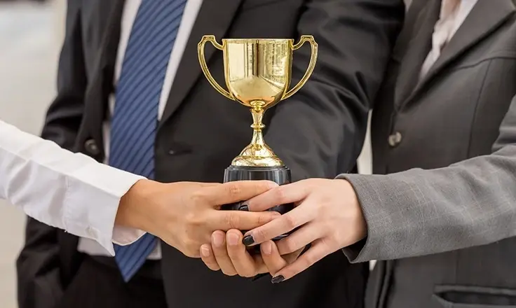 A cropped photo of three businesspeople, with distinct hands, collectively holding a golden trophy cup.