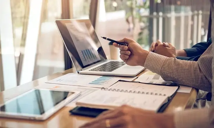 A professional using a pen to point at a bar chart on a laptop screen, with various printed reports and a notebook spread out on the desk.