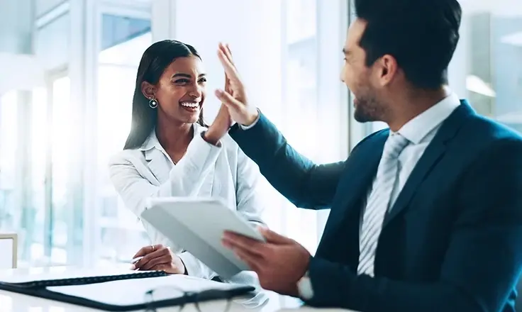 Two office workers in a bright, modern office, smiling and giving each other a high five.