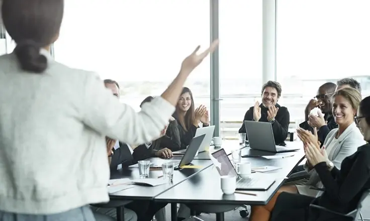 A group of professionals sitting around a conference table, smiling and clapping for a colleague giving a presentation.