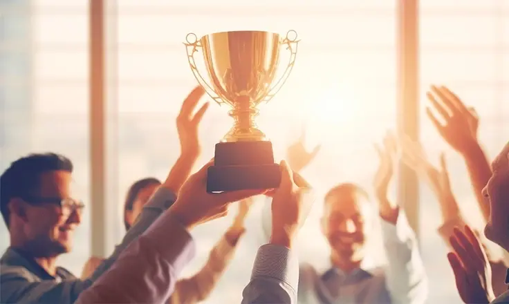 A group of cheerful business professionals celebrating a victory by holding up a gold trophy together in a sunlit office.