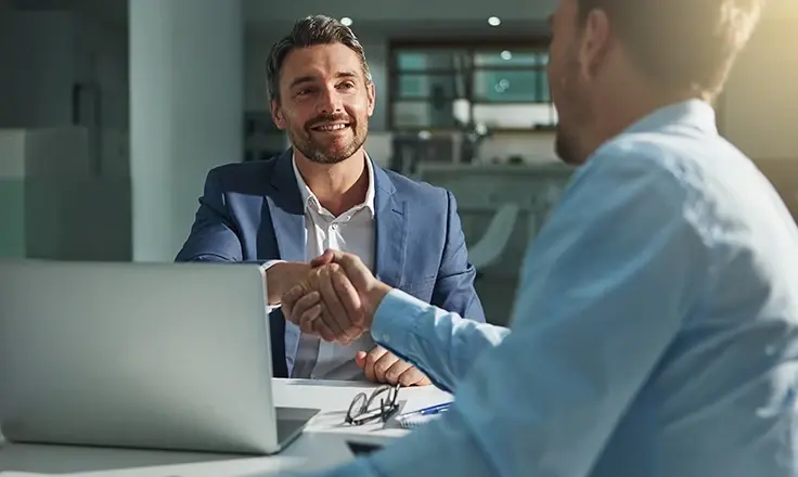 Two men in professional attire shaking hands across a desk in a modern office.