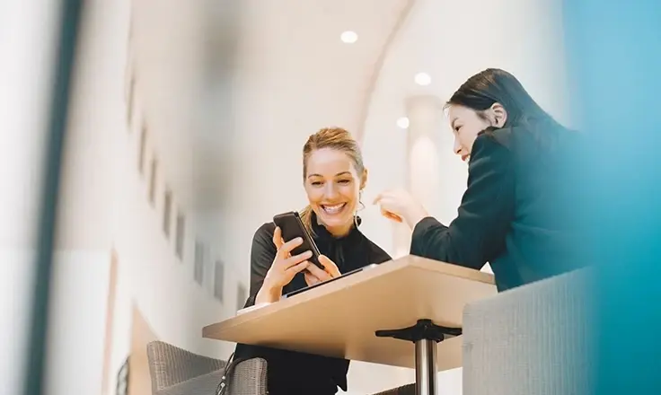 Two women sitting at a table in a bright, modern indoor space, smiling and looking at a smartphone together.