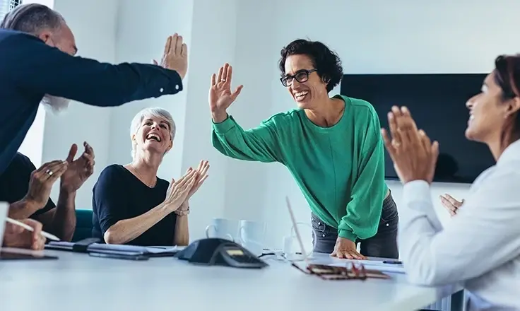 A group of professionals cheering and high-fiving during a celebratory meeting in a bright, modern office.