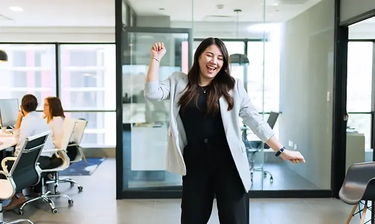 A young businesswoman in a light grey blazer cheering and dancing happily in the middle of a bright, modern office space.