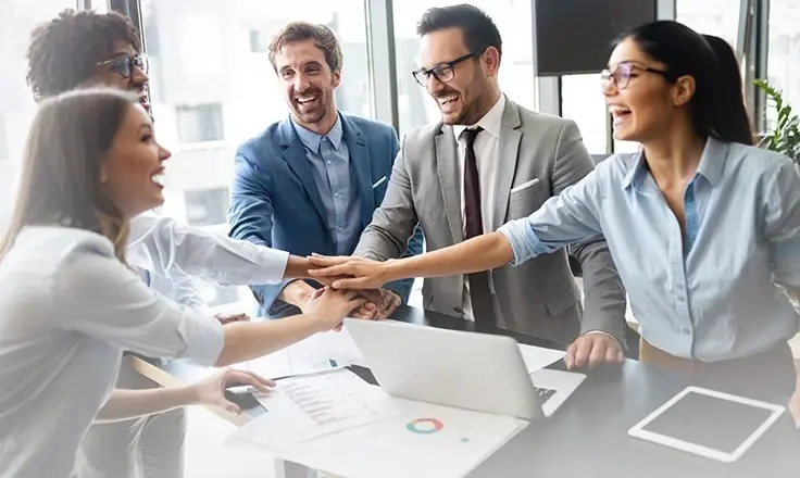 A group of coworkers laughing and putting their hands together in a huddle at a bright office desk.
