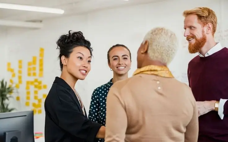 A multi-ethnic group of four colleagues talking and smiling in a modern office with sticky notes in the background.
