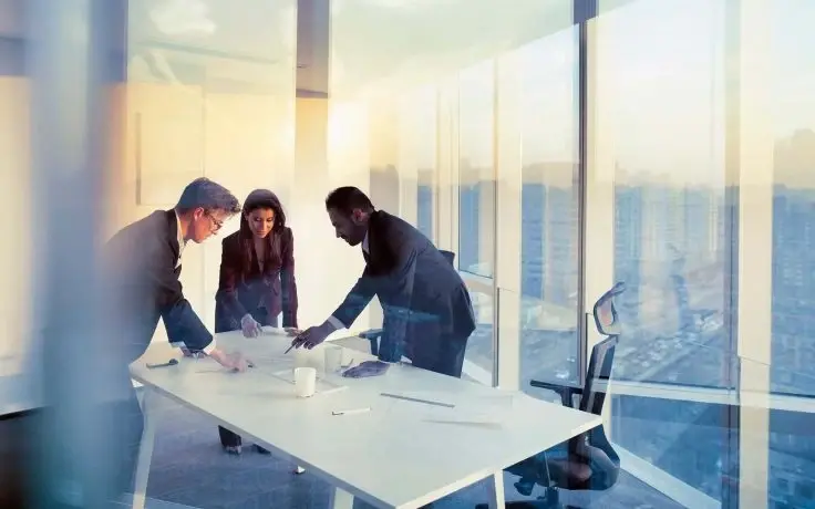 Three colleagues examine papers on a white table in a glass-walled office overlooking a city at sunset.
