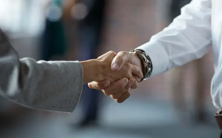A close-up of two people in business attire shaking hands, with one wearing multiple bracelets and a watch.