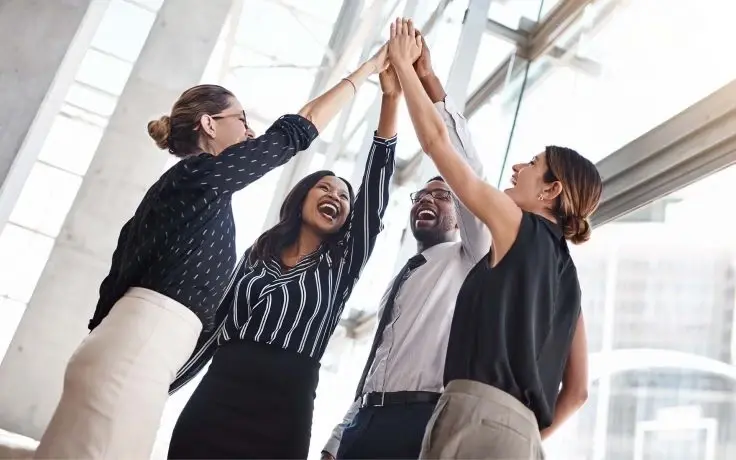 A business team of four, viewed from below, celebrates with a high five and laughter in a bright, modern office with windows.