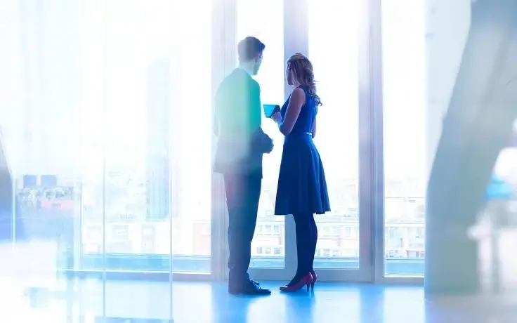 Two business professionals standing by a large floor-to-ceiling window in a bright office, looking at a tablet.