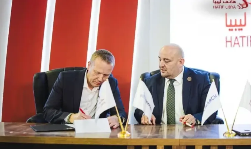 Two men in suits seated at a wooden desk, signing documents during an official ceremony for Hatif Libya and Comarch.