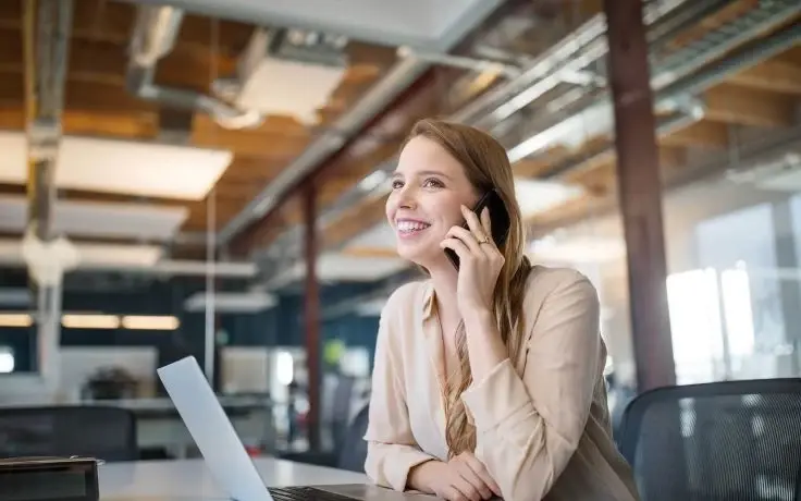 Smiling young woman talking on a smartphone while working on a laptop in a modern, open-plan office.