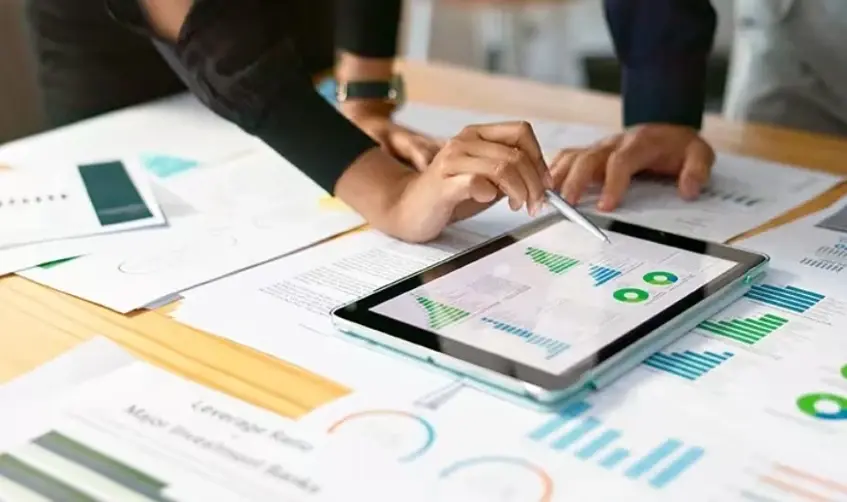 A woman pointing at green bar charts on a tablet screen with a stylus, surrounded by printed reports and graphs on a desk.