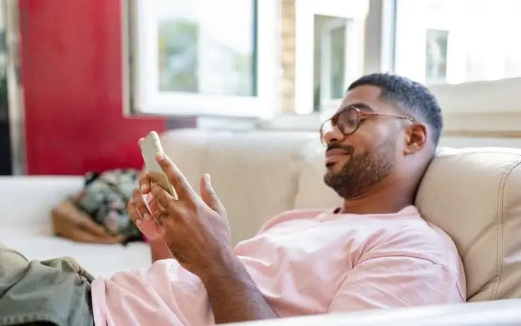 A relaxed young man with glasses and a pink shirt relaxing on a couch while scrolling through his smartphone.