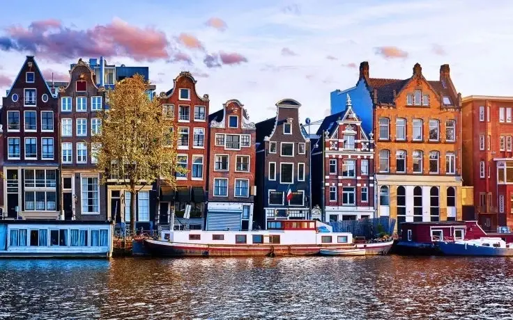 A scenic view of Amsterdam's historic gabled buildings along a canal, with several houseboats docked on the water.