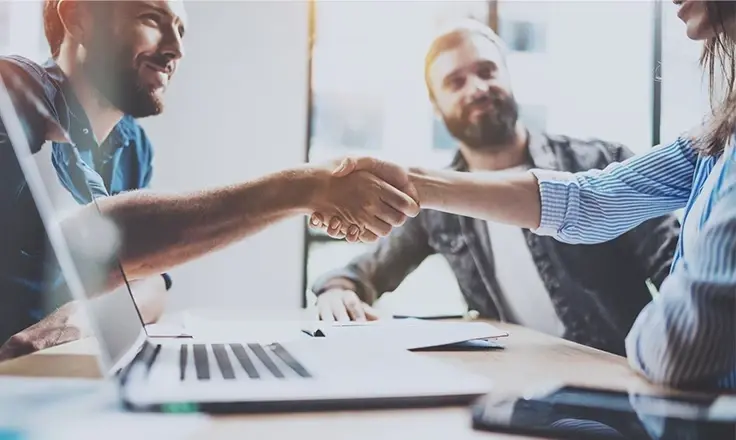 A man and a woman shake hands over a desk with a laptop, while another bearded man watches in a brightly lit office.