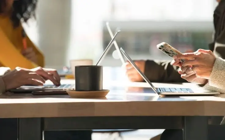 A close-up of hands using laptops and a smartphone at a wooden table with a black coffee mug in a sunlit room.