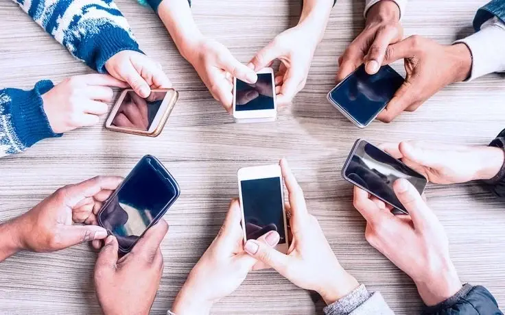 A top-down view of a group of people holding and using smartphones in a circle over a wooden table.