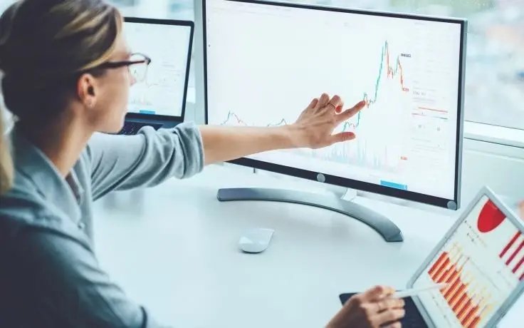 A person in glasses points to a rising stock market chart on a large monitor at a desk with a laptop and a tablet.