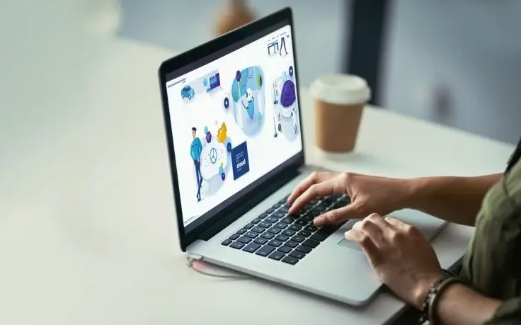 A person types on a laptop keyboard with a screen displaying various digital illustrations and icons.
