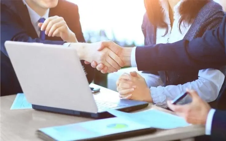 Two business professionals shake hands over a desk with a laptop and documents during a meeting.
