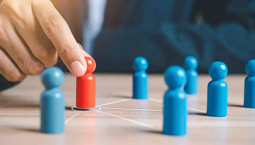 A close-up of a person's hand placing a single red wooden figure among several blue figures on a table.