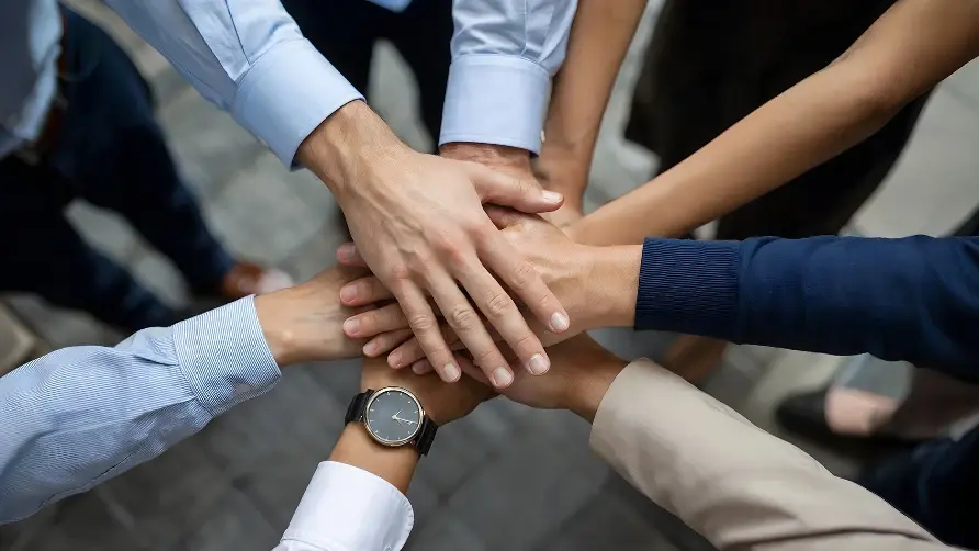 A top-down, close-up view of a diverse group of people in business attire joining their hands together in a central huddle.
