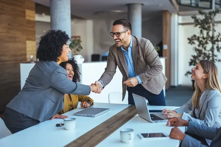 A group of smiling business professionals in a modern office, with a man and woman shaking hands across a conference table.