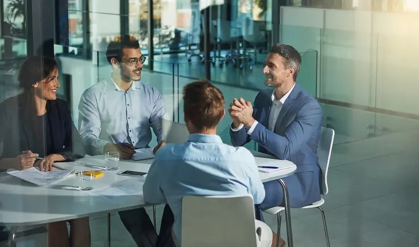 A team of diverse professionals discussing papers and charts during a meeting at a large conference table in a glass office.