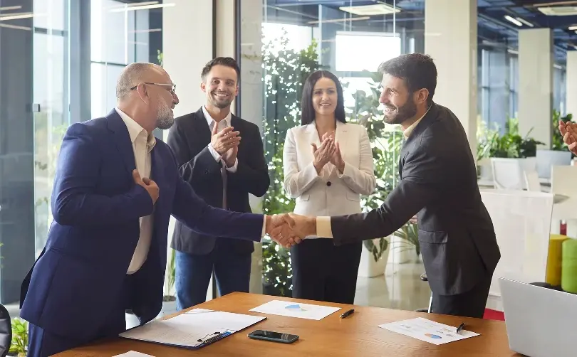 Two businessmen shake hands over a table while colleagues clap in a bright, modern office with large windows.