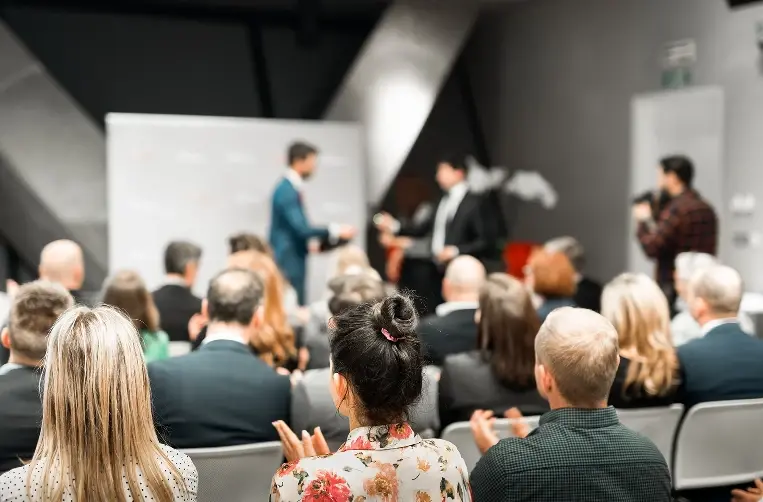An audience seated in a conference room, seen from behind, clapping for two speakers on stage during a presentation.