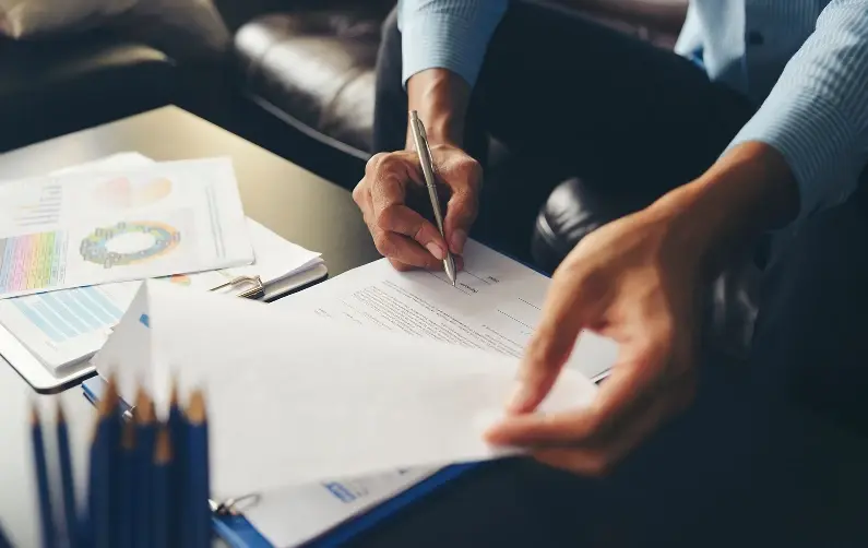 Close-up of a person signing a paper document while colleagues place hands on top of each other over charts on a desk.