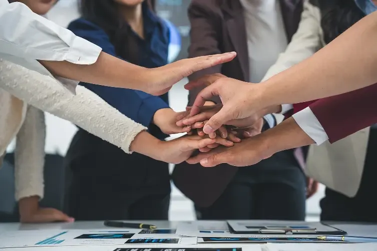 A group of colleagues joining their hands together in a central stack over a desk covered in financial reports and charts.