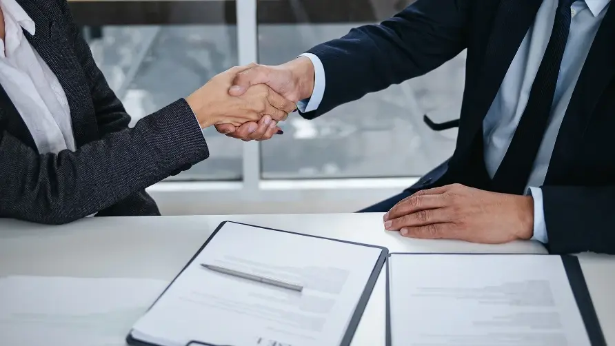 Two business professionals shaking hands across a white desk, with several documents and a pen laid out in front of them.
