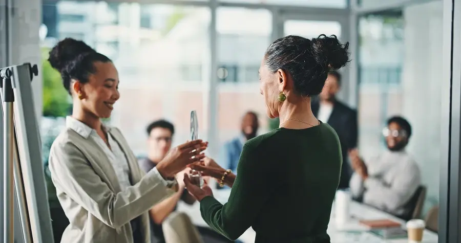A professional woman smiling as she receives a glass award in a bright, modern office setting with team members applauding in the background.